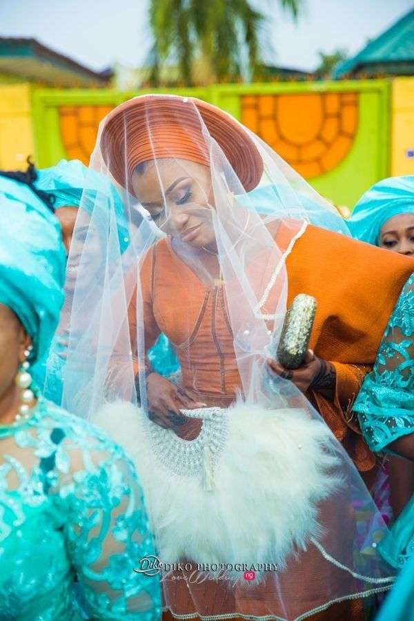 Yoruba bride in burnt orange Aso Oke and gele with a white veil, holding a feathered fan.