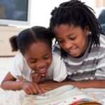 An older boy and a younger girl smile while reading a picture book on a white rug.
