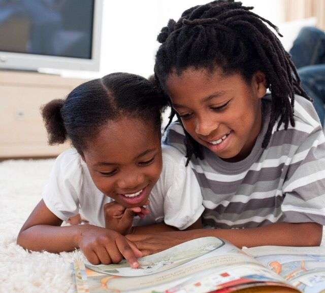 An older boy and a younger girl smile while reading a picture book on a white rug.
