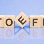 Wooden blocks spelling "TOEFL" on a reflective blue surface, with the letter "E" tilted.